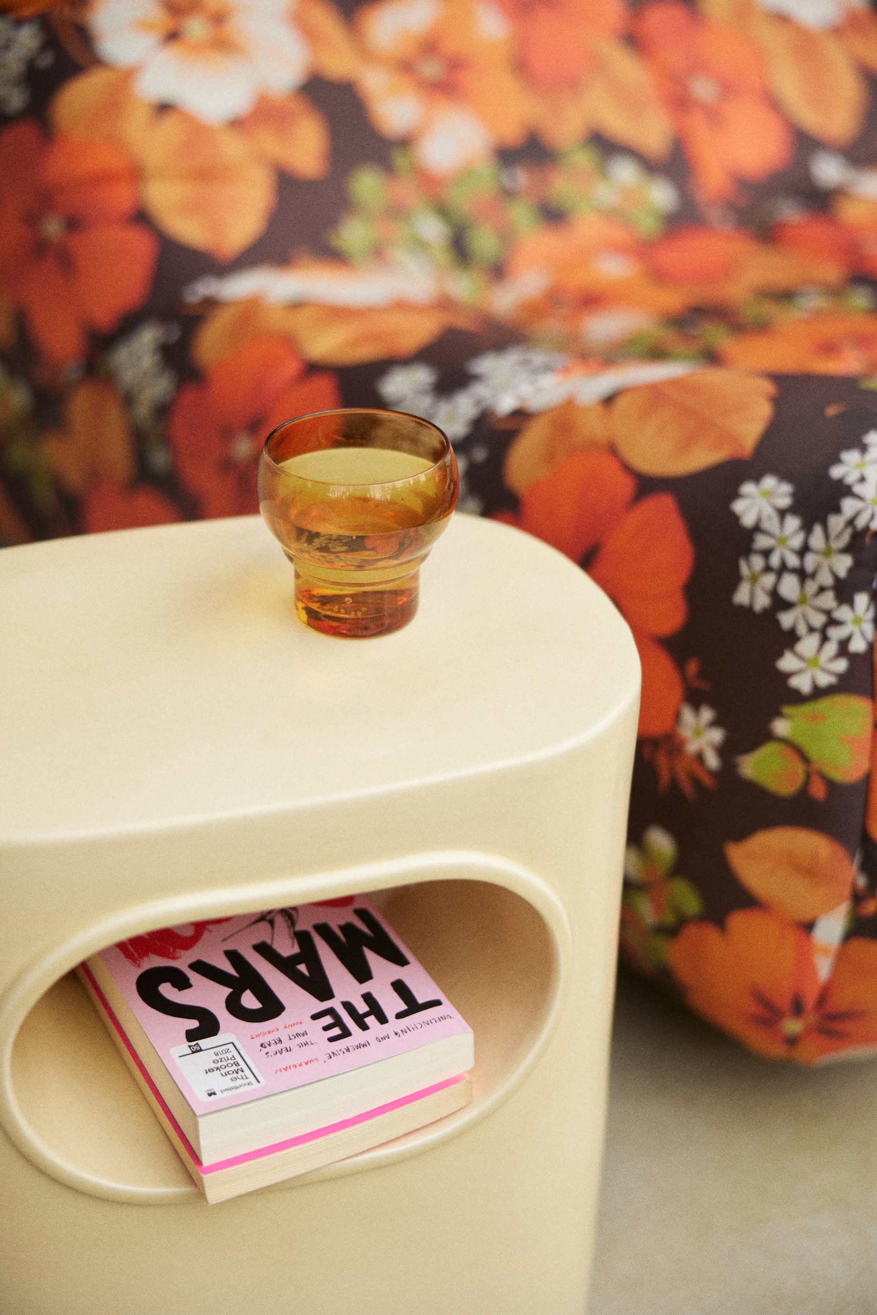 A small beige table with a circular cutout holds a pink book titled “THE MARS…” and an amber HKLIVING 70s Glassware Bulb Glass, Set of 4. In the background, there’s a sofa with a bold orange floral pattern.