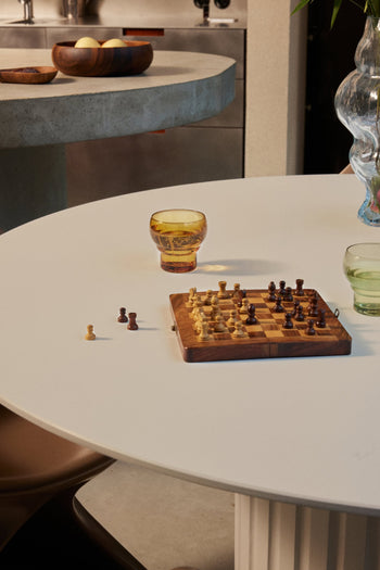 A small chessboard with pieces rests on a round white table next to HKLIVING 70s Glassware Bulb Glasses—one amber and one green. A few chess pieces are off the board, with a fruit bowl in the background.