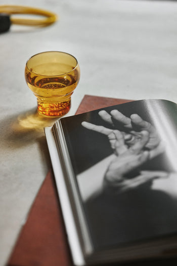 An amber drink in an HKLIVING 70s Glassware Bulb Glass rests beside an open book with a black and white photo of moving hands, all on a light surface.
