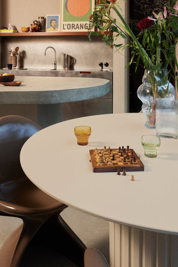 A round white table with a chess set and two HKLIVING 70s Glassware Bulb Glasses sits in a modern kitchen. In the background are a sink, vase of flowers, and a framed L’Orange poster on the counter.