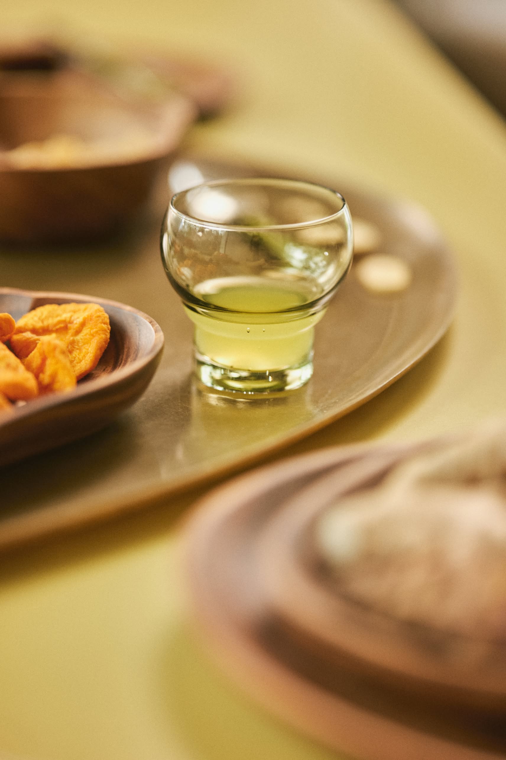 An HKLIVING 70s Bulb Glass, filled with light green liquid, rests on a wooden tray beside a dish of orange snacks, all arranged on a pale green surface.