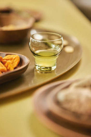 An HKLIVING 70s Bulb Glass, filled with light green liquid, rests on a wooden tray beside a dish of orange snacks, all arranged on a pale green surface.