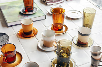 HKLIVING 70s Glassware: Coffee Cups, Set of 4 by HKLIVING are arranged with matching saucers on a white tiled tabletop, beside a closed book and folded cloth napkins in the background.
