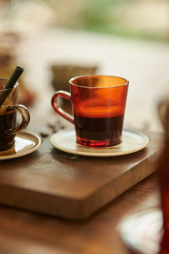 A red coffee cup from the HKLIVING 70s Glassware: Coffee Cups, Set of 4 sits on a white saucer atop wood, filled with dark coffee, with another cup and blurred background elements in view.