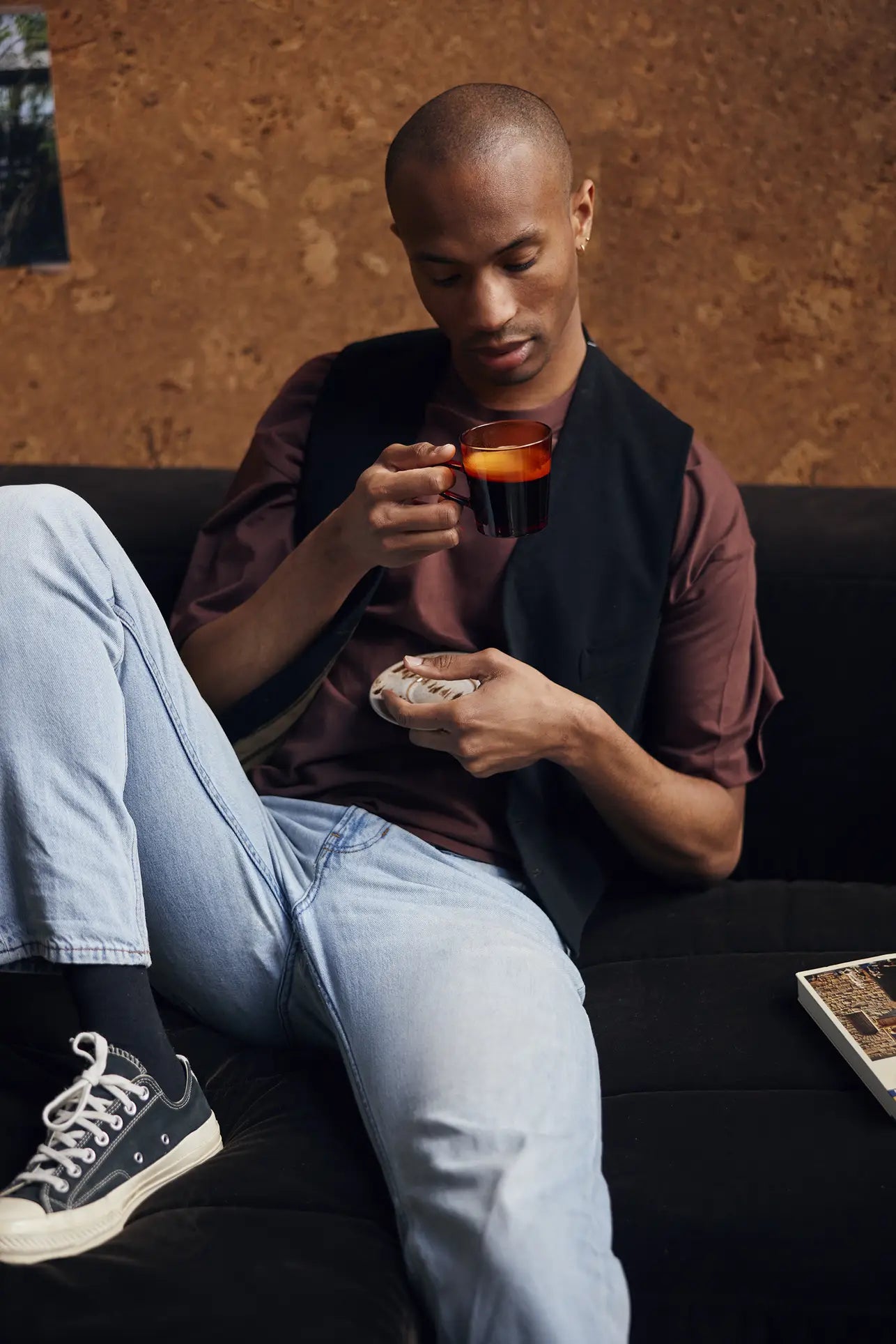 A person sits on a black sofa, holding an HKLIVING 70s Glassware Coffee Cup and saucer, wearing a brown shirt, light blue jeans, black vest, and black Converse sneakers. A book rests nearby.
