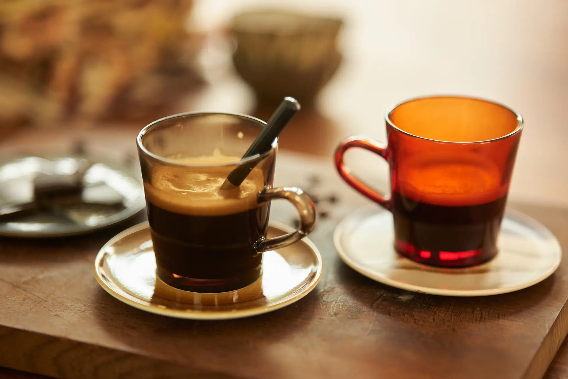 Two cups from the HKLIVING 70s Glassware Coffee Cups Set sit on saucers atop wood; one amber and empty, the other clear with coffee and a spoon. A blurred plate of chocolates appears in the background.