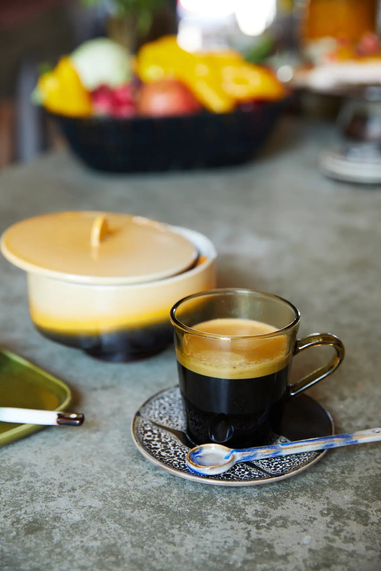 A HKLIVING 70s Glassware coffee cup sits on a saucer with a spoon, while an HKLIVING lidded sugar bowl and a blurred fruit basket rest in the background, all atop a gray countertop.