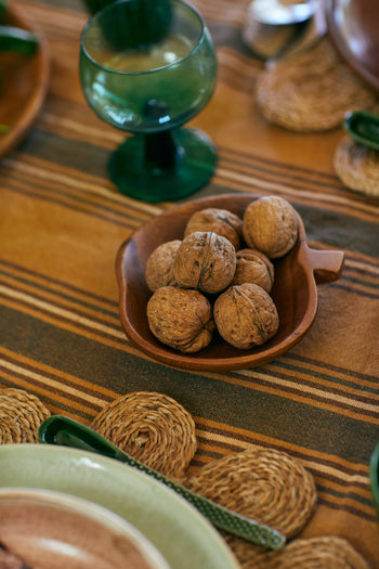 An HKLIVING Acacia Apple Bowl M filled with whole walnuts is set on a striped brown tablecloth, surrounded by green glassware, ceramic plates, and woven placemats on a styled dining table.