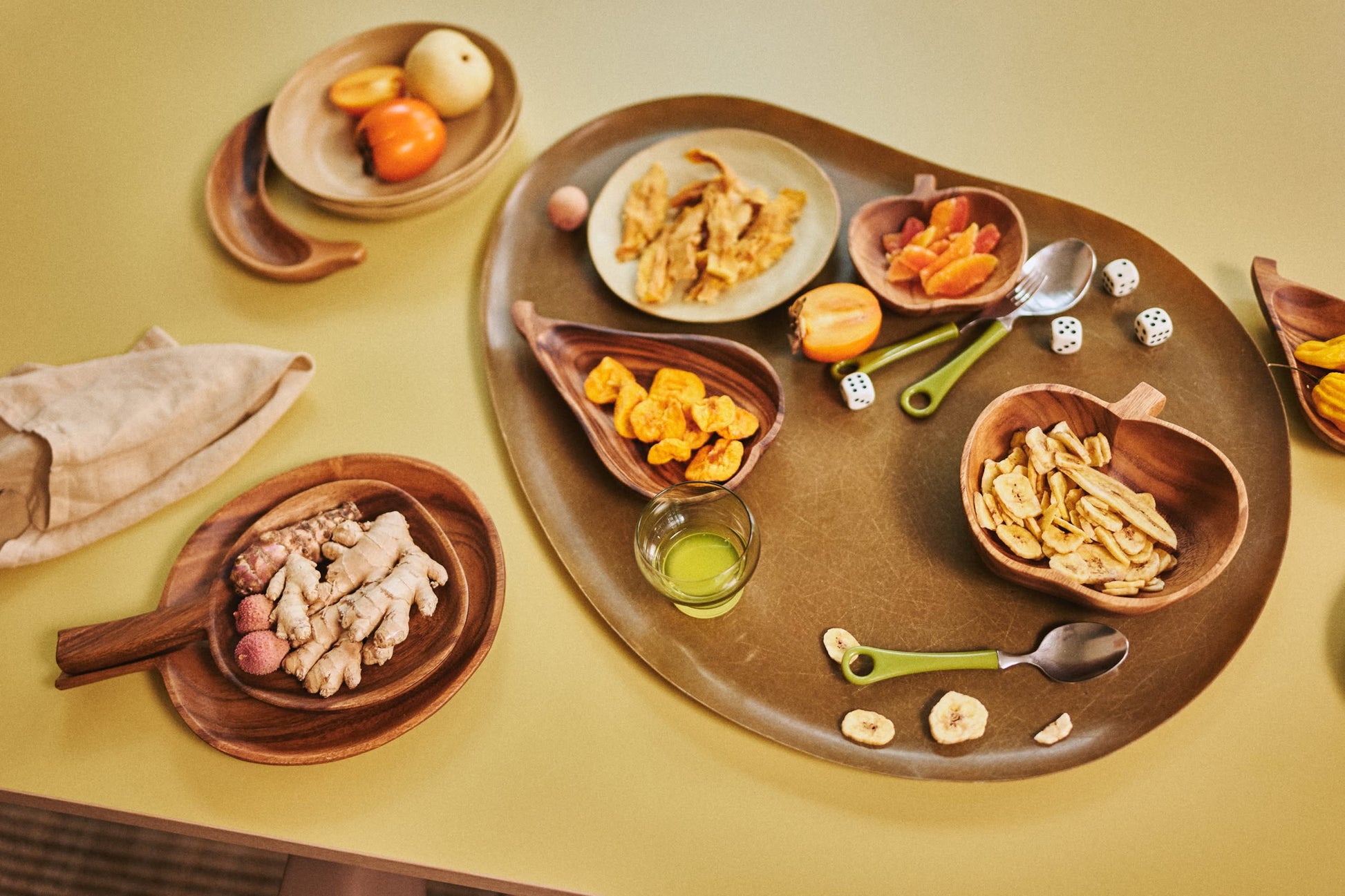 A tabletop displays assorted wooden bowls with dried fruits, ginger, whole fruits, a green drink, dice, and metal spoons, all arranged on the HKLIVING Acacia Platter L by HKLIVING. A napkin rests beside the organic-shaped platter.