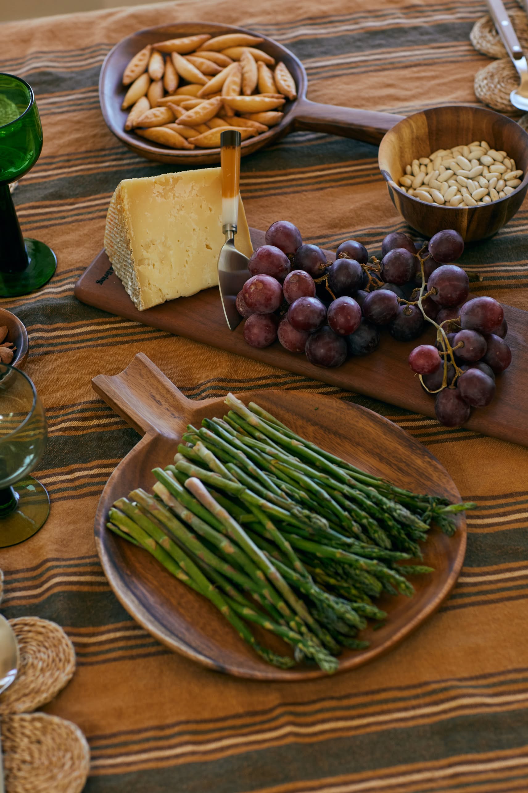 An HKLIVING Acacia Platter L displayed with asparagus, grapes, cheese and knife, crackers, and pine nuts sits atop a striped tablecloth.