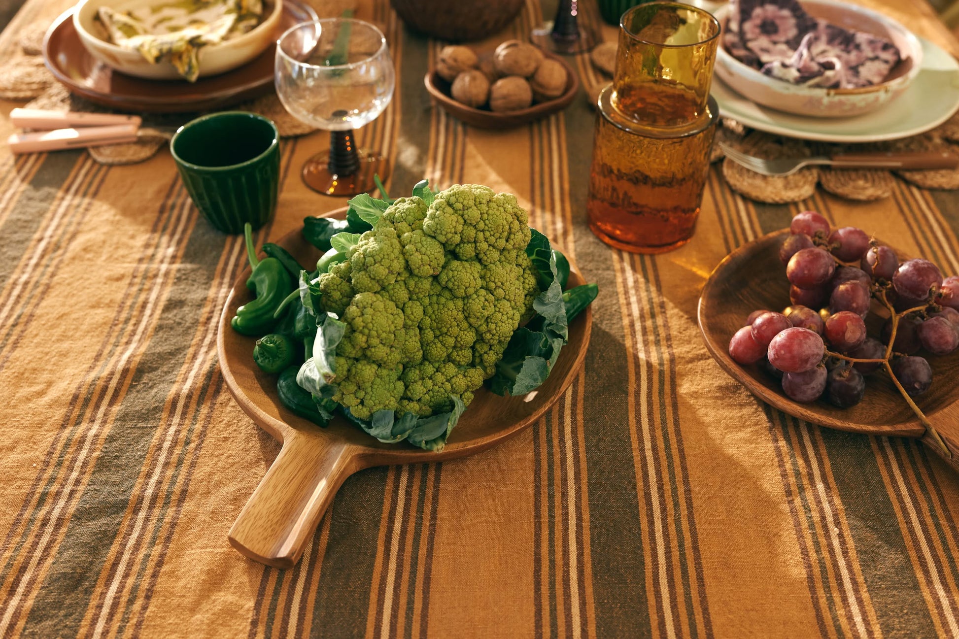 A wooden table set for a meal displays the HKLIVING Acacia Platter L by HKLIVING with a head of green cauliflower and leafy greens, surrounded by glasses, red grapes, walnuts, and more atop a striped tablecloth.