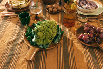 A wooden table set for a meal displays the HKLIVING Acacia Platter L by HKLIVING with a head of green cauliflower and leafy greens, surrounded by glasses, red grapes, walnuts, and more atop a striped tablecloth.