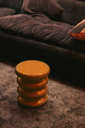 The HKLIVING Allure Side Table, a round orange piece with a stacked ribbed design, stands on a gray carpet in front of a dark plush sofa topped with a tan pillow.