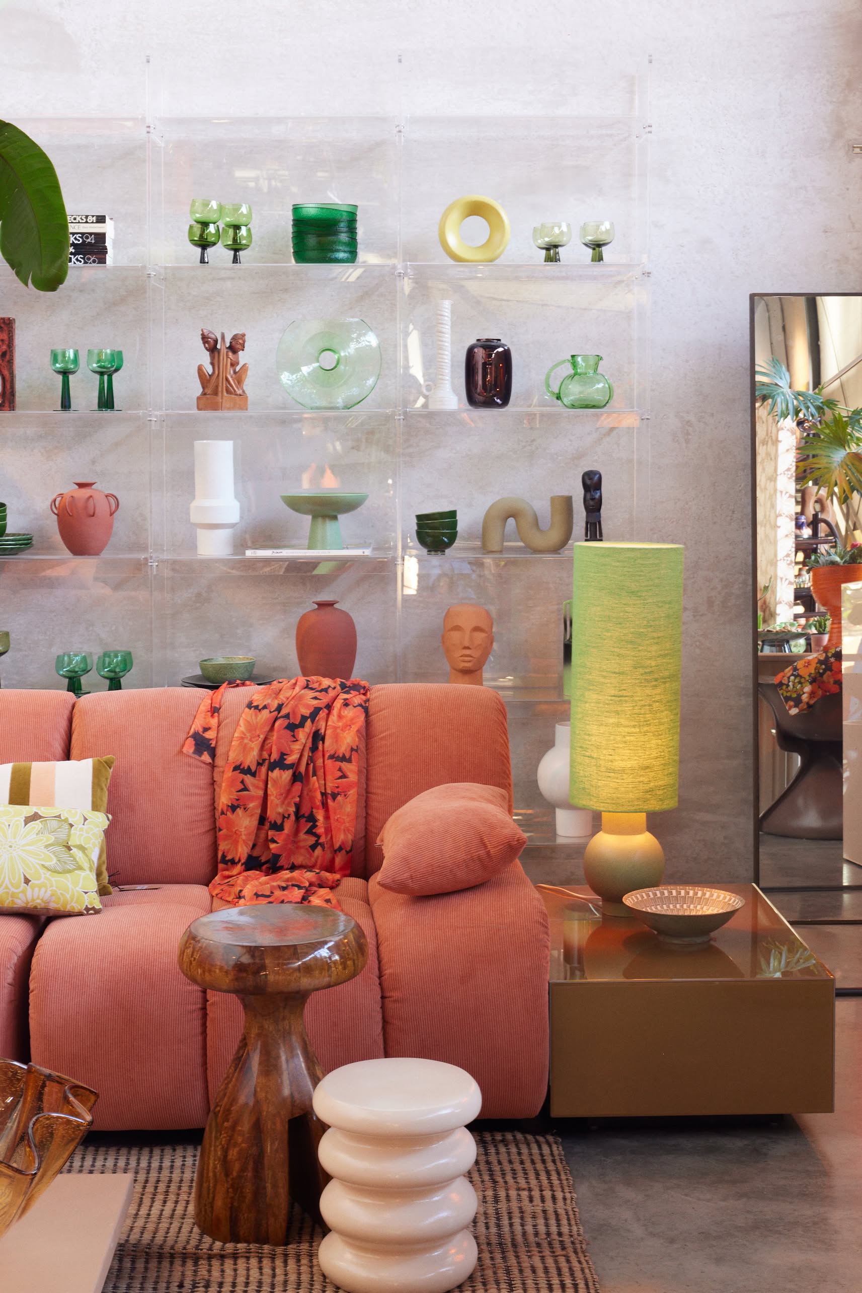A modern living room with a pink sofa, green lamp on the HKLIVING Allure Side Table, wooden and white ribbed stools, and a clear shelving unit displaying colorful vases. A mirror reflects plants and modern elegance in the background.