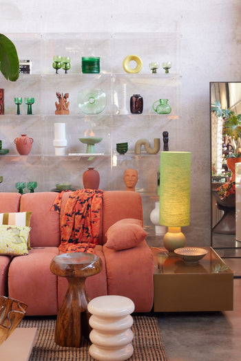 A modern living room with a pink sofa, green lamp on the HKLIVING Allure Side Table, wooden and white ribbed stools, and a clear shelving unit displaying colorful vases. A mirror reflects plants and modern elegance in the background.