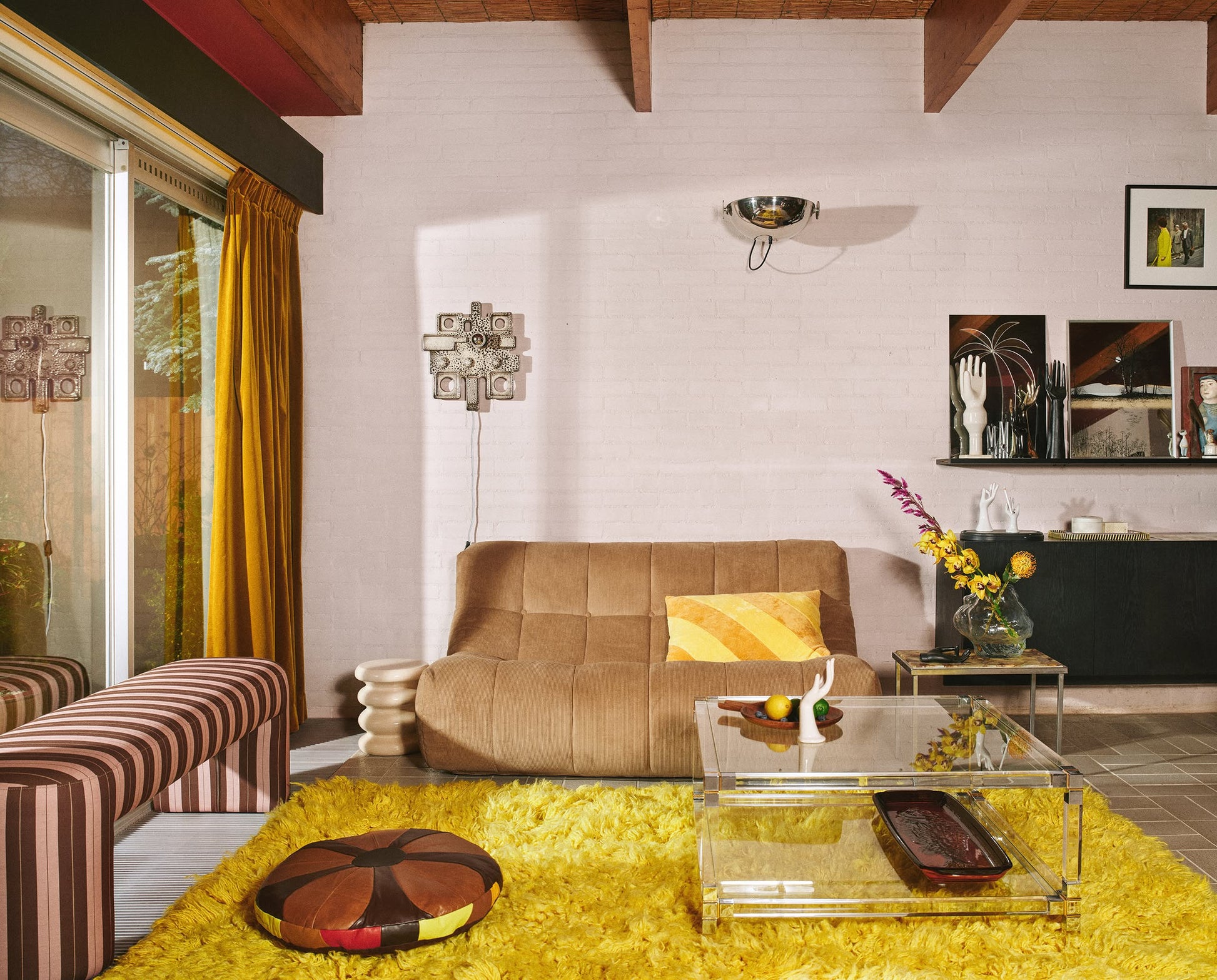 A retro living room with a tan loveseat, yellow shag rug, striped ottoman, HKLIVING Allure Side Table by HKLIVING, glass coffee table, mustard yellow curtains, eclectic wall art, and wooden beams illuminated by sunlight.