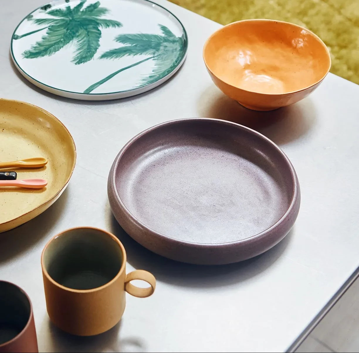A set of HKLIVING Bold & Basic Ceramics Deep Plates in two colors—brown and deep yellow—displayed stylishly on white, alongside a vibrant purple bowl, beige mug, and a plate with a green palm tree design.