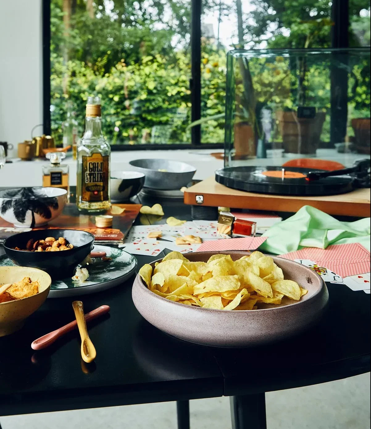 A tabletop with stylish tableware, HKLIVING Bold & Basic Ceramics Deep Plate (Set of 2) filled with potato chips, snacks, playing cards, Gold Strike liquor, and a record player in a bright room with large windows overlooking greenery.