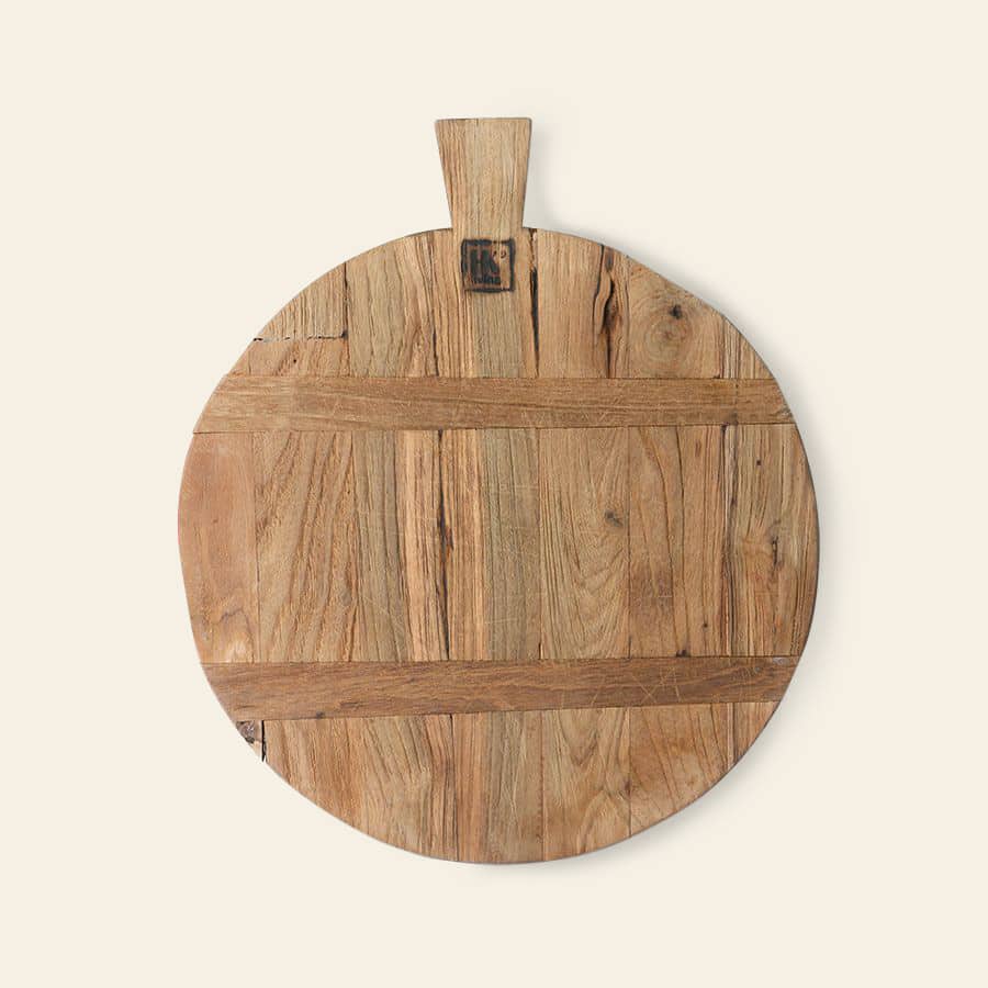 The HKliving Bread Board Reclaimed Teak, Medium, by HKliving, features a round shape with a short handle. Crafted from reclaimed teak, its unique wood grain and natural imperfections offer rustic charm against a light backdrop.