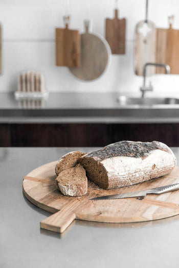 A rustic loaf with two slices cut rests on the HKliving Bread Board Reclaimed Teak, Medium by HKliving, accompanied by a bread knife in a modern kitchen, with wooden boards hanging in the background for added warmth.