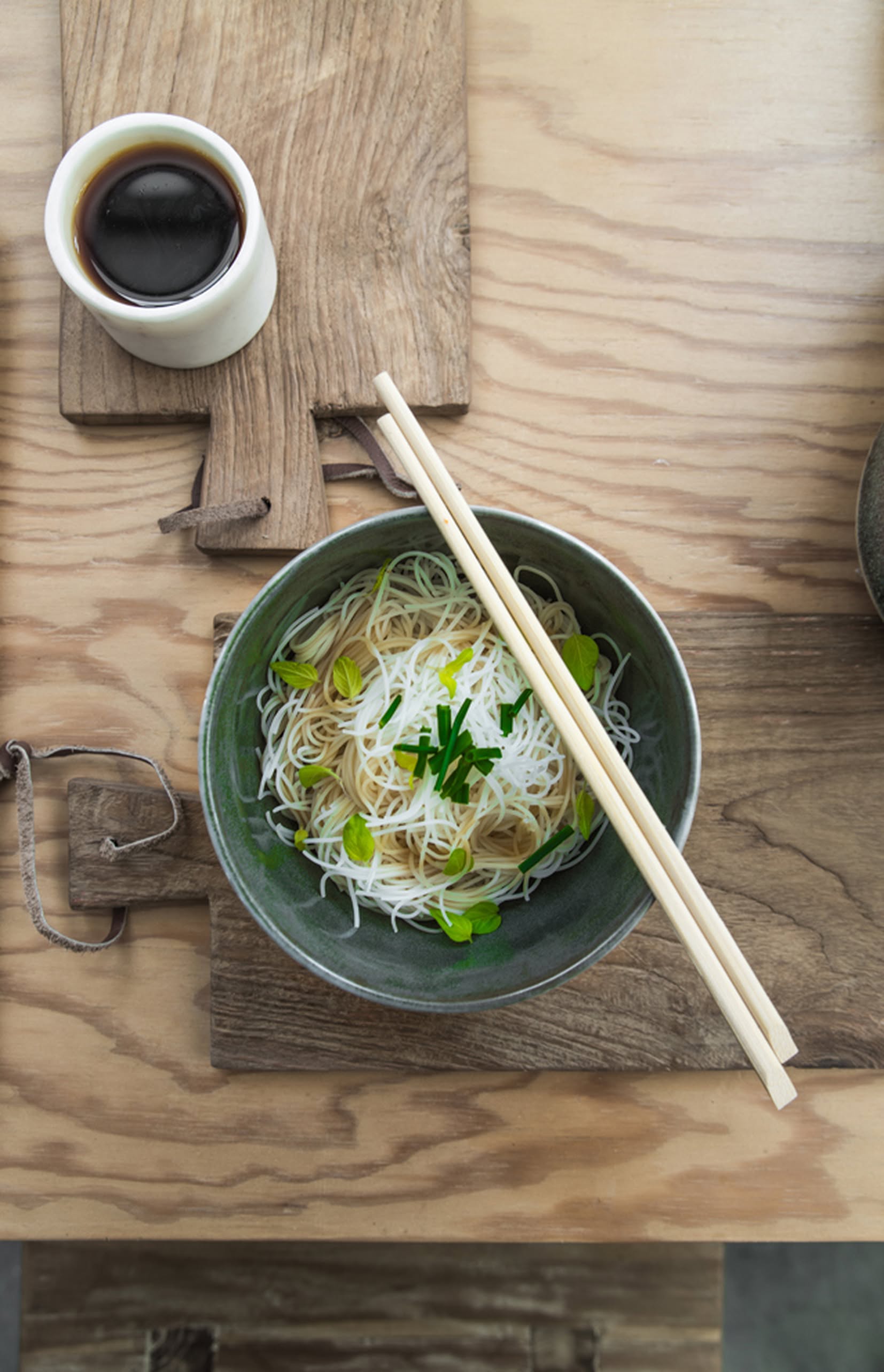 A bowl of noodles topped with green onions and herbs, served with chopsticks and soy sauce on rustic HKLIVING Bread Board Teak, Set of 3 by HKLIVING, arranged on a wooden table.