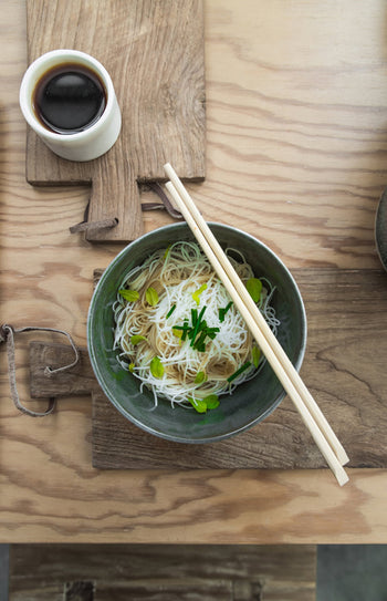 A bowl of noodles topped with green onions and herbs, served with chopsticks and soy sauce on rustic HKLIVING Bread Board Teak, Set of 3 by HKLIVING, arranged on a wooden table.