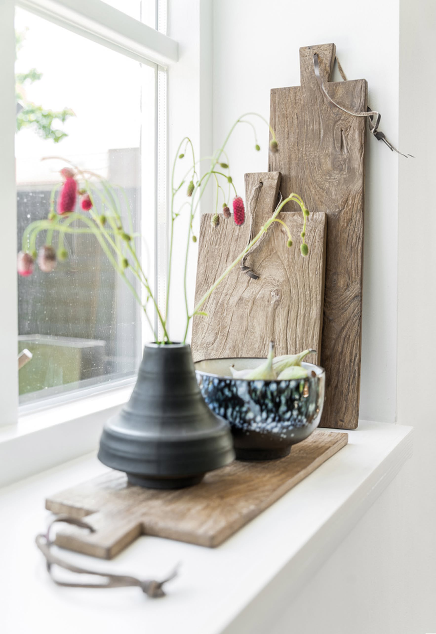 On the windowsill, a dark ceramic vase with pink flowers, a blue patterned bowl, and several rustic teak boards—including the HKLIVING Bread Board Teak, Set of 3—by HKLIVING are arranged upright as sunlight pours in through the window.