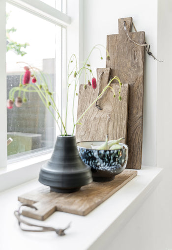On the windowsill, a dark ceramic vase with pink flowers, a blue patterned bowl, and several rustic teak boards—including the HKLIVING Bread Board Teak, Set of 3—by HKLIVING are arranged upright as sunlight pours in through the window.