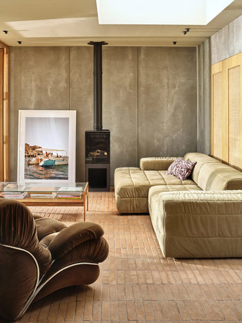A modern living room features the HKLIVING Brut Sofa, Left by HKLIVING with sleek curves, a plush brown chair, glass coffee table, black wood stove, and a framed boat photo against a textured gray wall on brick flooring.