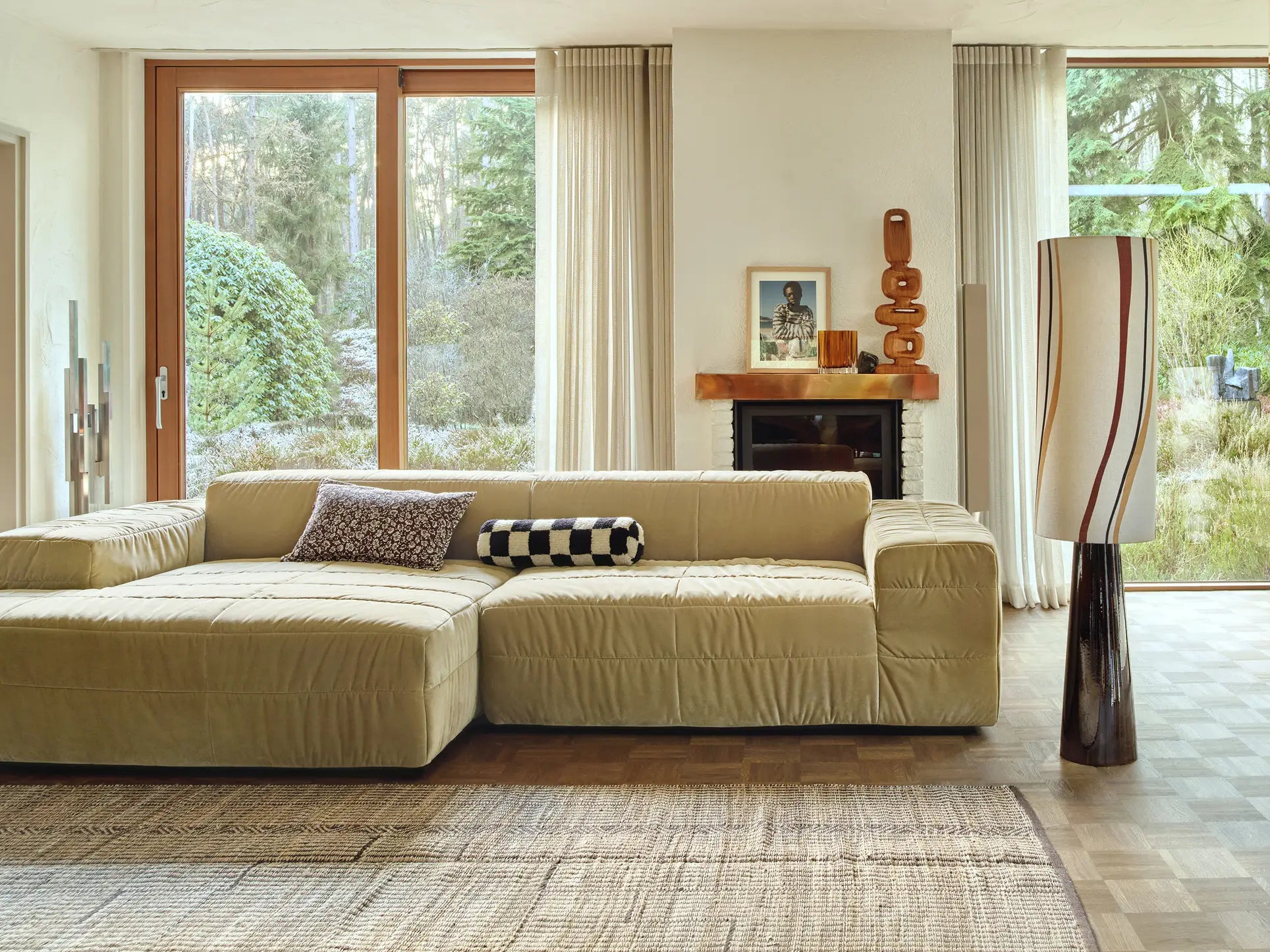 A modern living room featuring the HKLIVING Brut Sofa, Right Divan, by HKLIVING, with patterned cushions, floor-to-ceiling garden-view windows, a decorated fireplace mantel, sheer curtains, and a tall sculptural floor lamp.