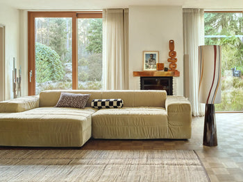 A modern living room featuring the HKLIVING Brut Sofa, Right Divan, by HKLIVING, with patterned cushions, floor-to-ceiling garden-view windows, a decorated fireplace mantel, sheer curtains, and a tall sculptural floor lamp.