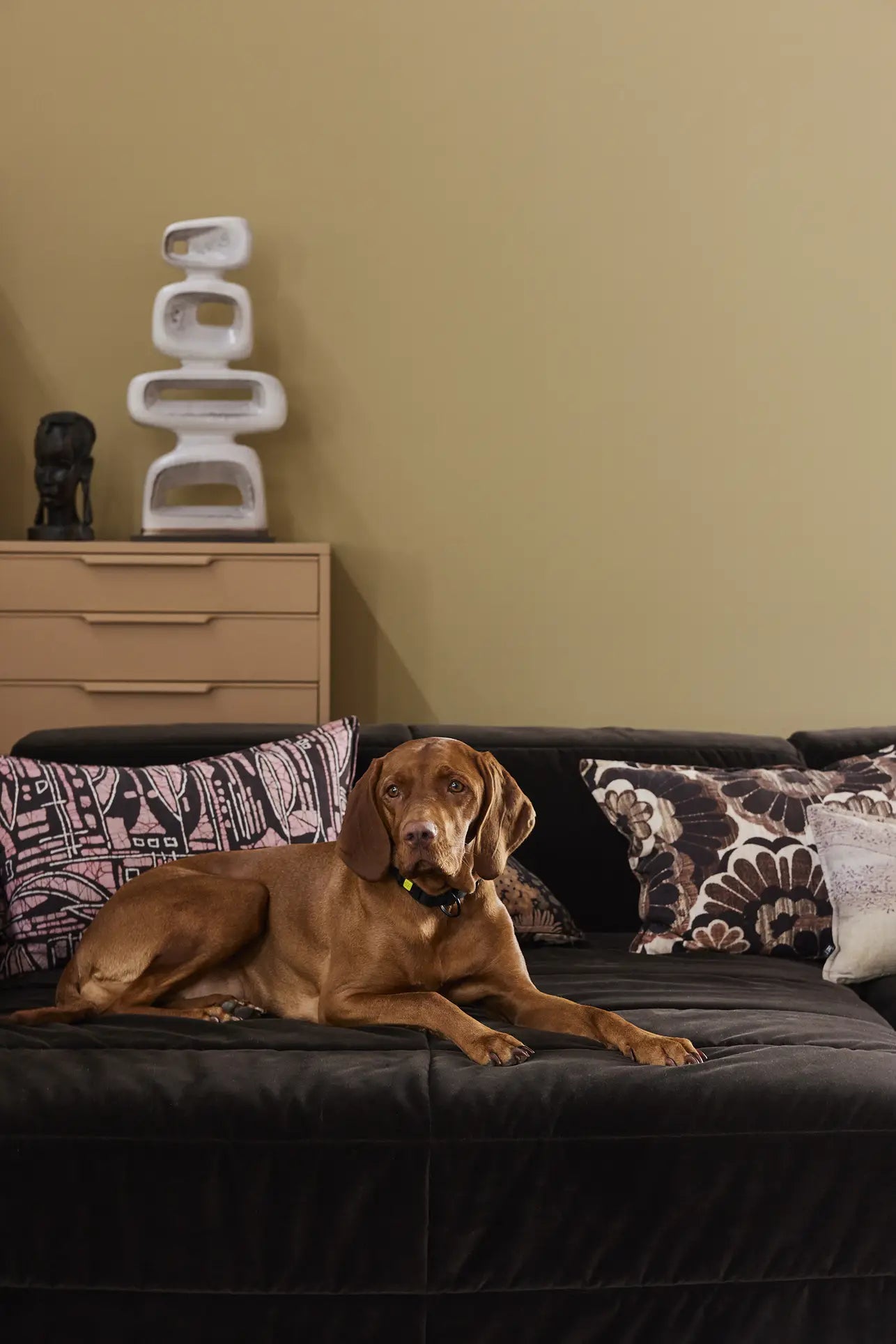 A brown dog rests on the HKLIVING Brut Sofa, Right Divan, with patterned cushions in a modern living room. Behind the sofa are beige drawers, decorative vases, and minimalist sculpture pieces by HKLIVING.