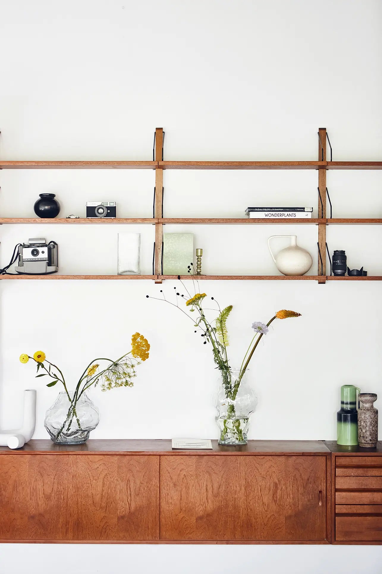 A wooden shelf with vintage cameras, books, and decor sits above a cabinet. Two glass vases with wildflowers and the HKLIVING Ceramic Jar M by HKLIVING add charm to this minimalist space inspired by Mediterranean interiors.
