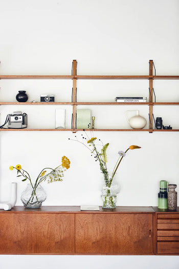 A wooden shelf with vintage cameras, books, and decor sits above a cabinet. Two glass vases with wildflowers and the HKLIVING Ceramic Jar M by HKLIVING add charm to this minimalist space inspired by Mediterranean interiors.