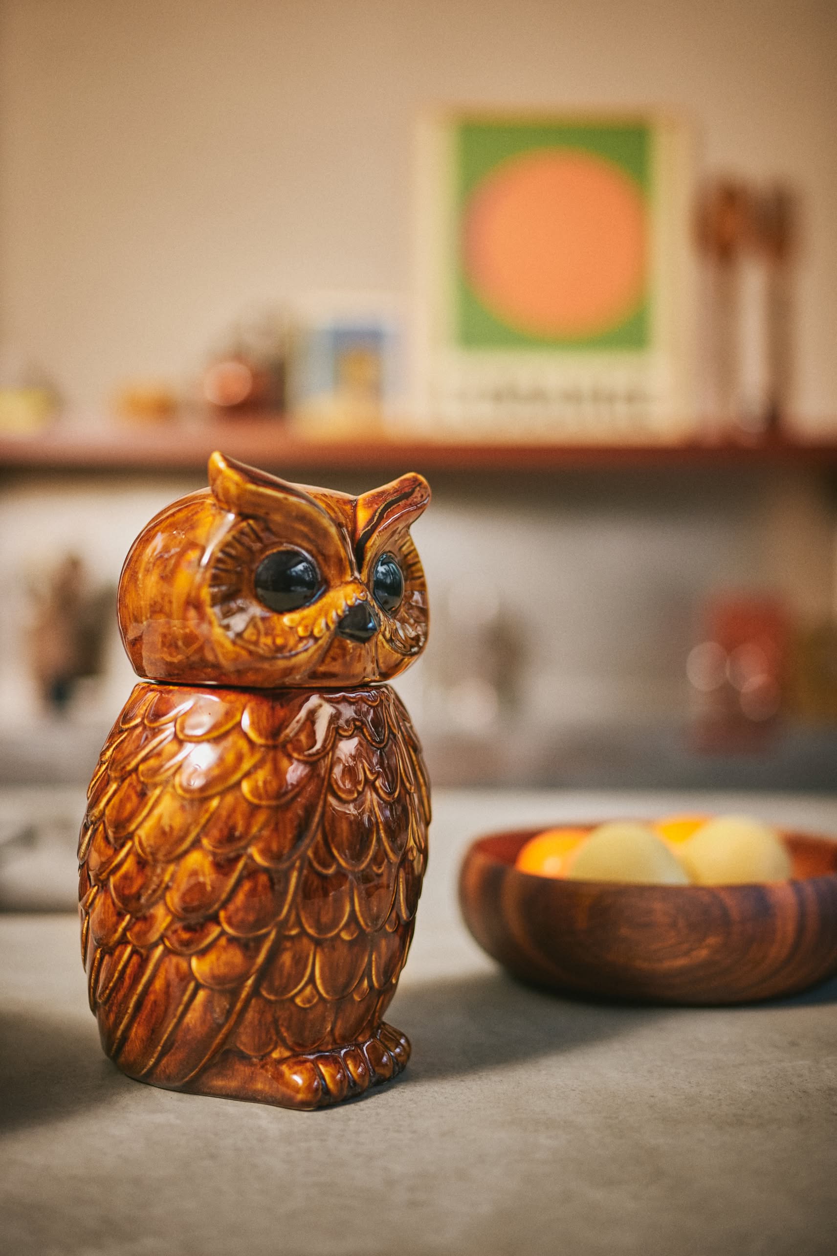 The HKLIVING Ceramic Owl Jar by HKLIVING, featuring detailed brown and orange feathers, sits on a countertop beside a wooden bowl of yellow objects, with a blurred, artfully decorated background.