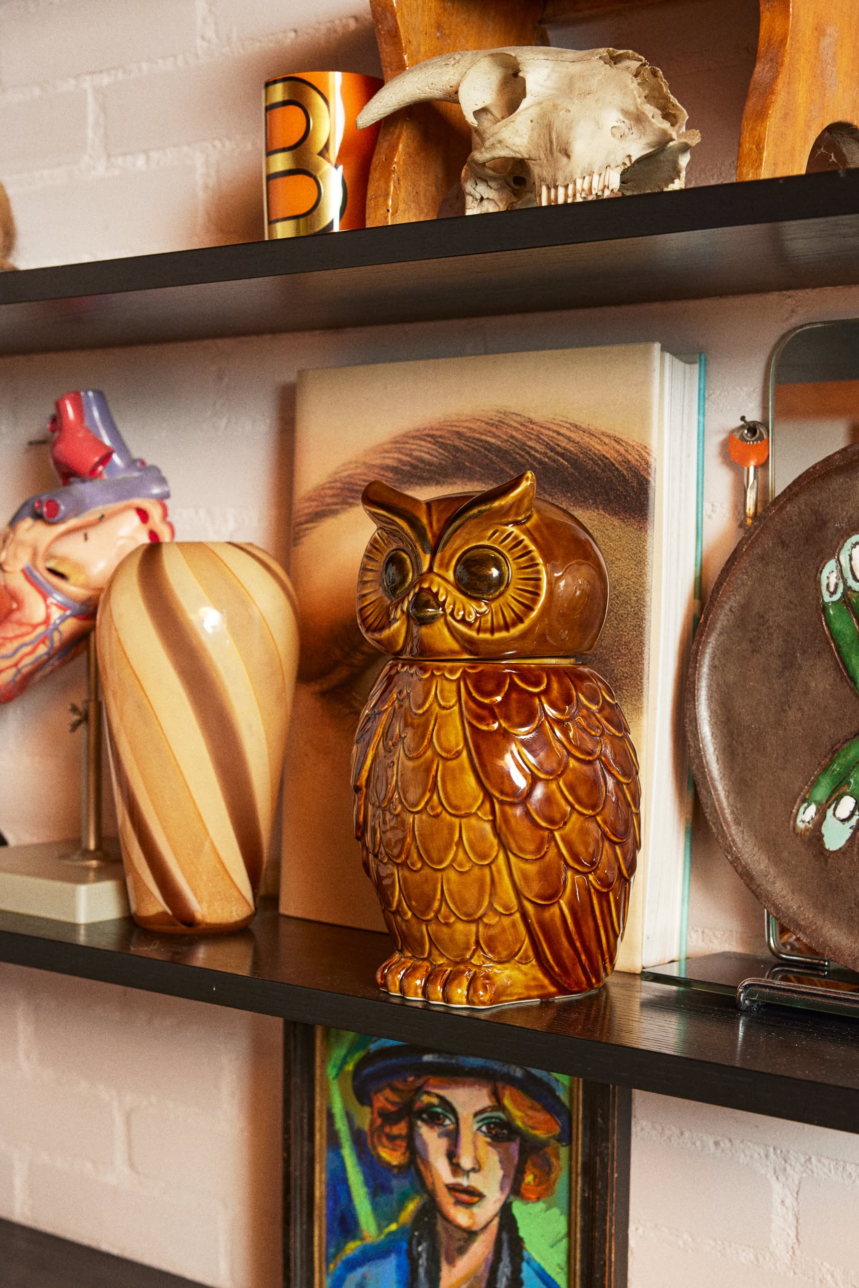 The HKLIVING Ceramic Owl Jar by HKLIVING rests on a shelf among eclectic decor: a striped vase, an eye-printed book, a skull, and colorful art—adding the perfect vintage accent to your space.