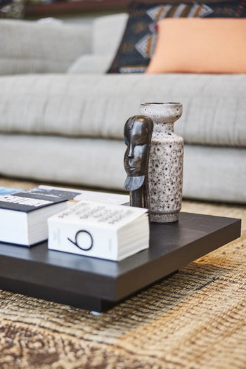 A close-up of a modern coffee table with books, an HKLIVING Ceramic Retro Vase, and a small dark sculpture. In the background is a beige sofa with a patterned cushion and a light brown pillow.