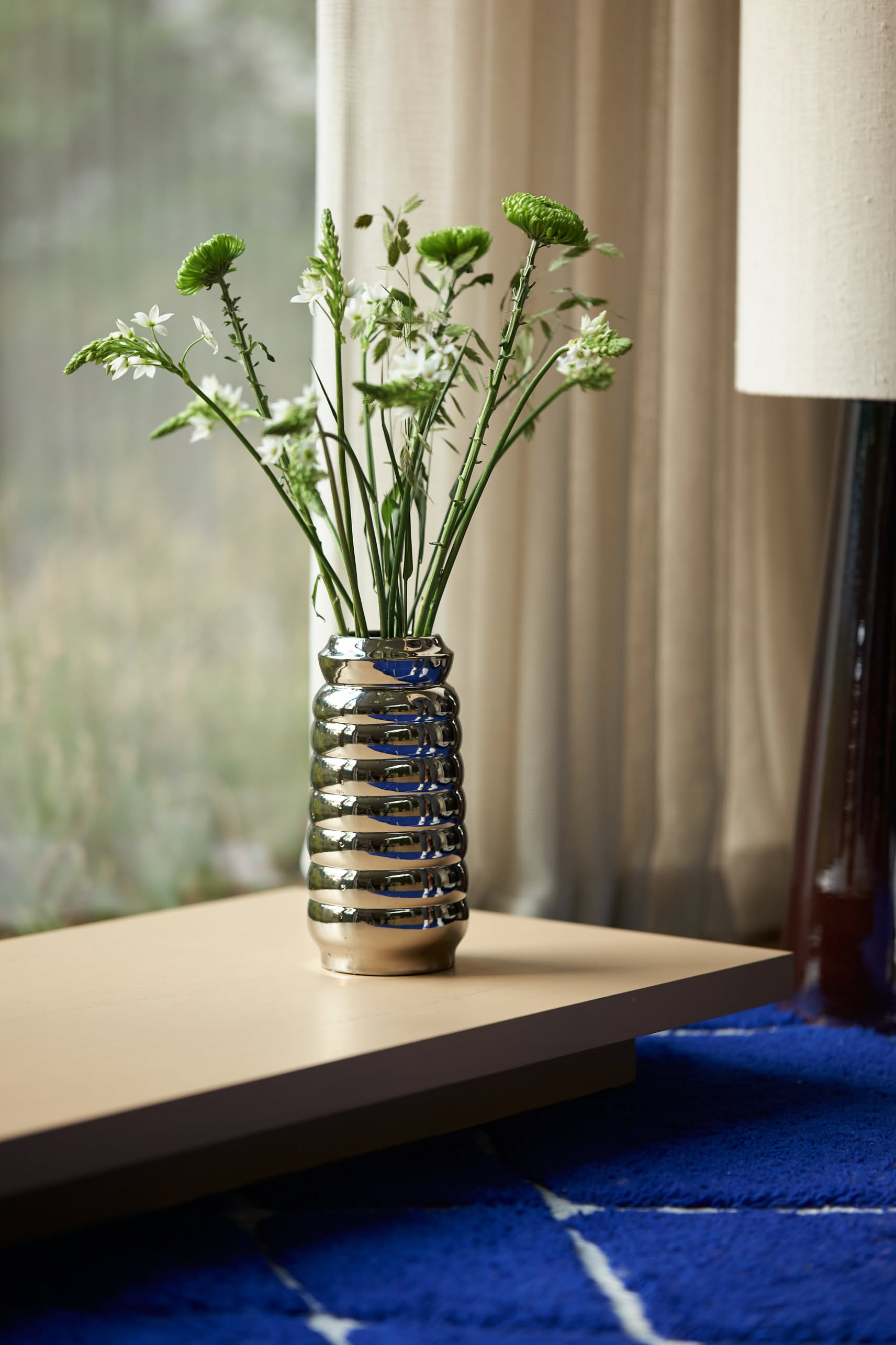 The HKLIVING Ceramic Ribble Vase with green and white flowers sits on a light wooden table by a blue rug, sunlight streaming through sheer curtains—ideal for modern interiors looking for a chic, reflective finish.