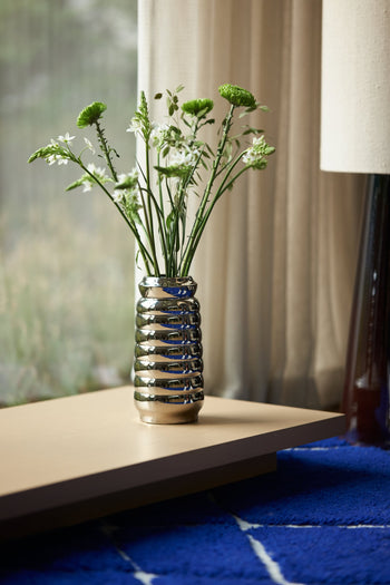 The HKLIVING Ceramic Ribble Vase with green and white flowers sits on a light wooden table by a blue rug, sunlight streaming through sheer curtains—ideal for modern interiors looking for a chic, reflective finish.