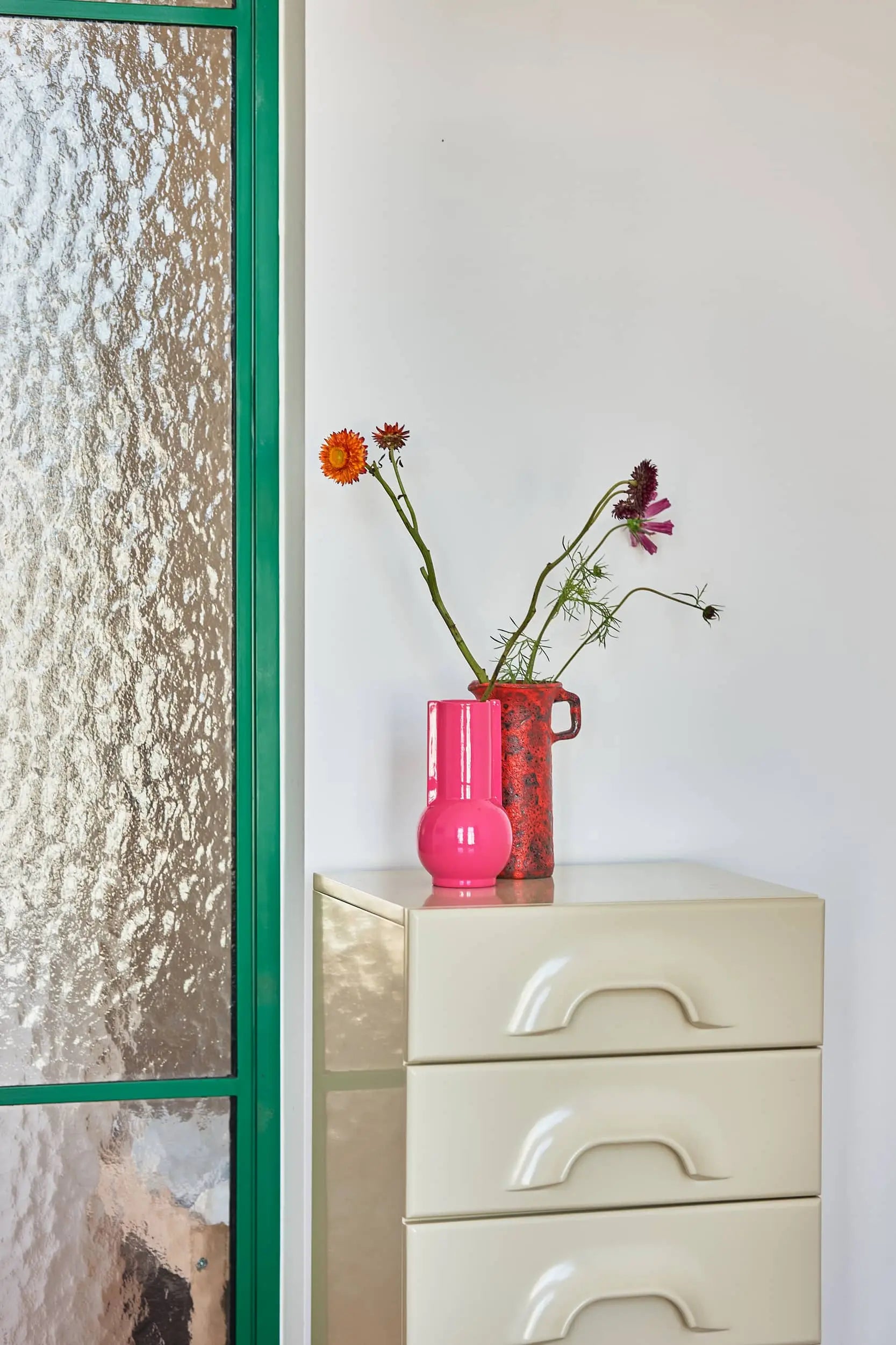 A beige chest of drawers with pink accents—featuring the HKLIVING Ceramic Vase Hot Pink and a red jug of flowers—stands by a textured glass door with a green frame, set against a white wall.