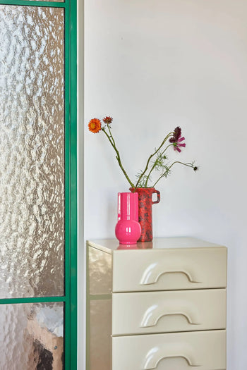 A beige chest of drawers with pink accents—featuring the HKLIVING Ceramic Vase Hot Pink and a red jug of flowers—stands by a textured glass door with a green frame, set against a white wall.