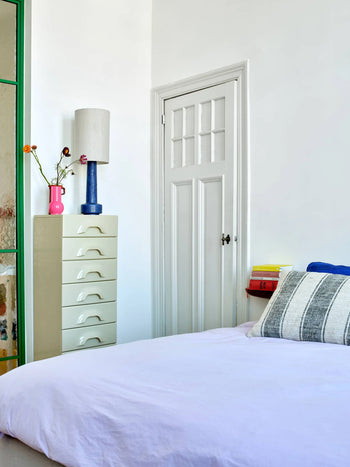 A bright bedroom with a bed covered in a light purple sheet, a striped pillow, a cream-colored dresser topped with a blue lamp and the HKLIVING Ceramic Vase Hot Pink by HKLIVING, and a small stack of books beside the bed.