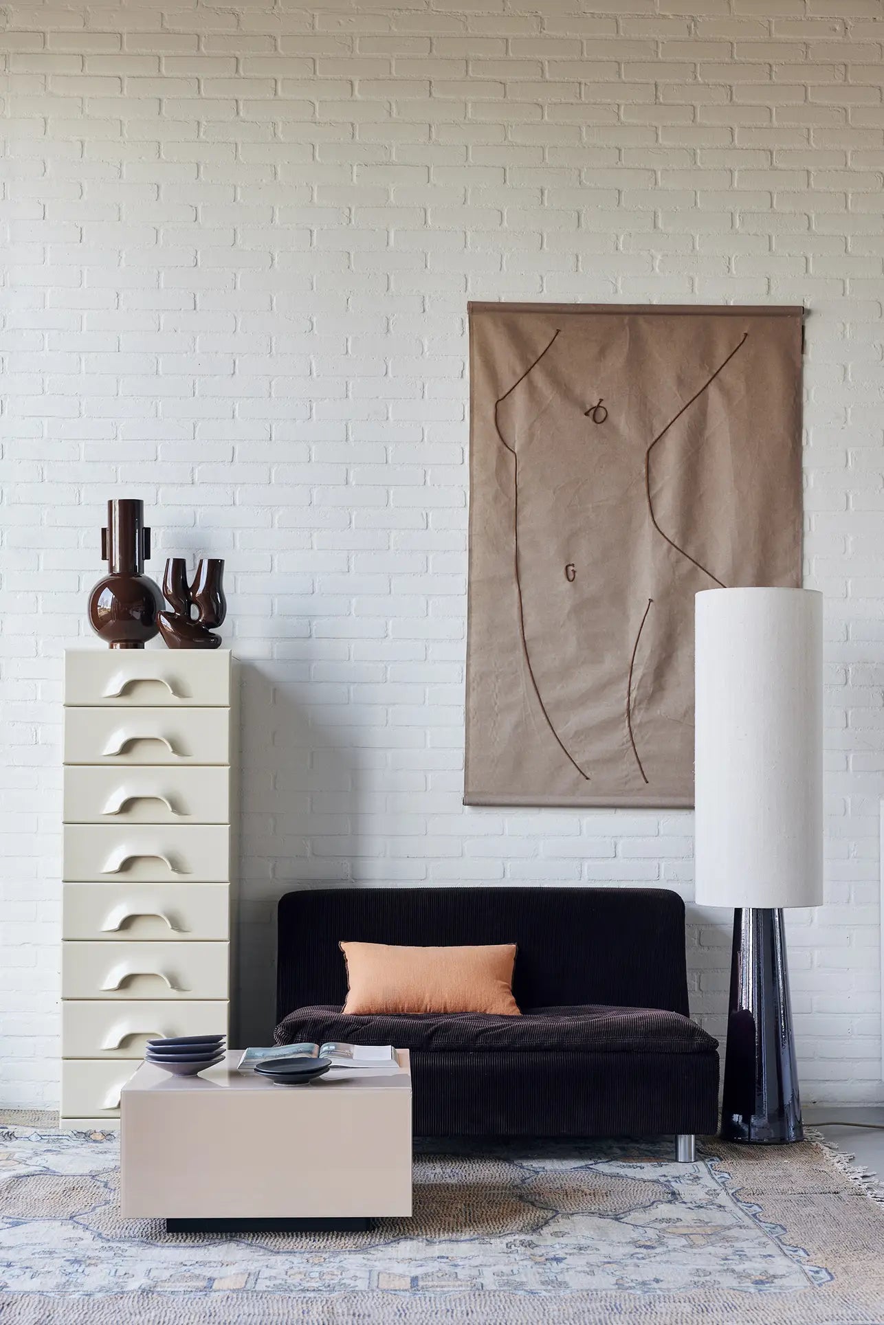 A modern living room with a black sofa, peach pillow, white cabinet with wavy drawers, tall lamp, abstract wall art, the HKLIVING Ceramic Vase L by HKLIVING on display, and a low white coffee table on a patterned rug against a white brick wall.