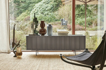 A modern living room displays the HKLIVING Ceramic Vase L by HKLIVING on a gray sideboard with decorative pieces, set atop a woven rug near large windows overlooking a garden. A black lounge chair appears partially in the foreground.