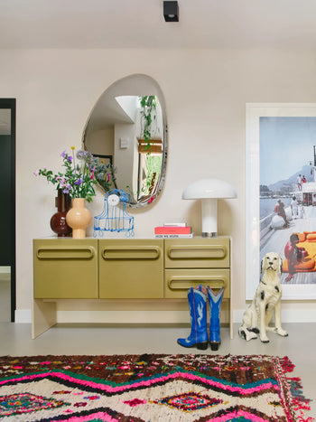 A modern entryway with a green sideboard, abstract mirror, HKLIVING Ceramic Vase L by HKLIVING with flowers, books, blue lamp, blue cowboy boots, dog statue, colorful rug, and a framed boat scene photo on the right wall.