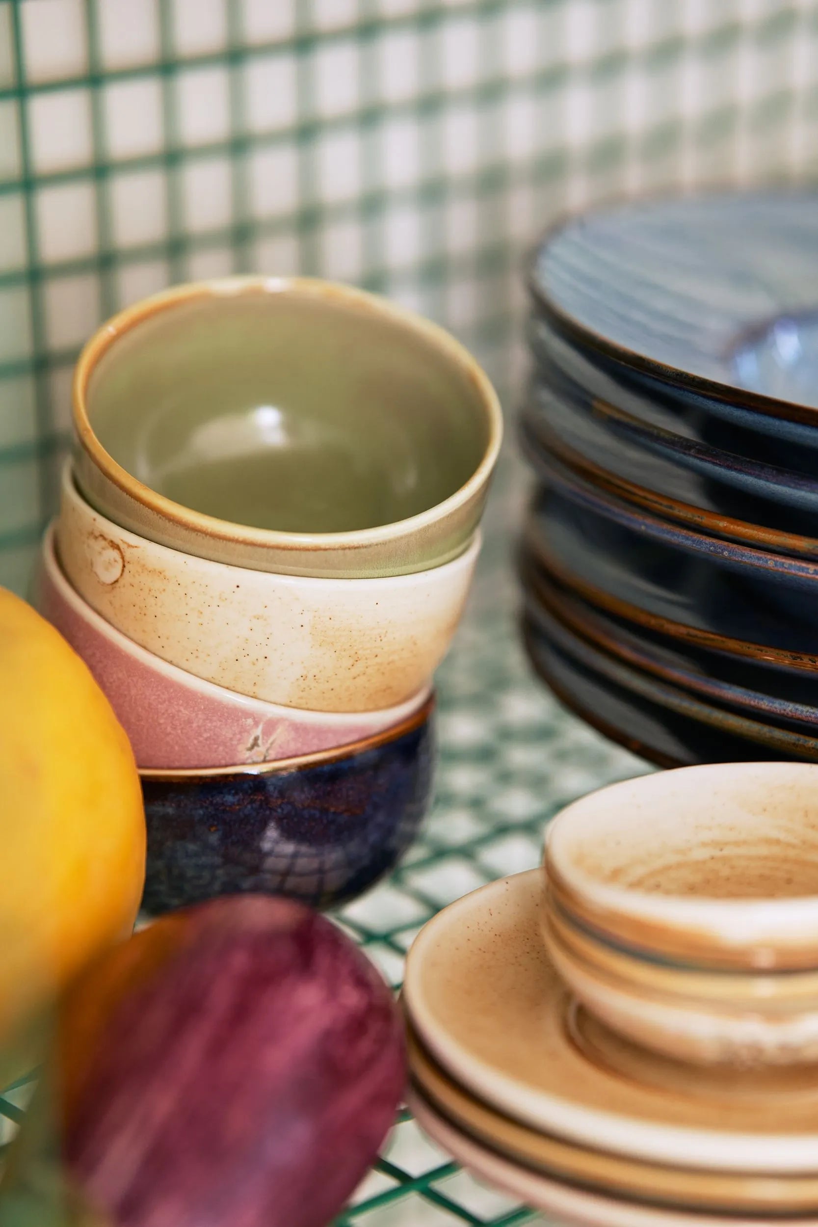 A close-up of HKLIVING Chef Ceramics Bowls in earthy tones, stacked on a grid-patterned surface, with a yellow lemon and a purple fig in the foreground.