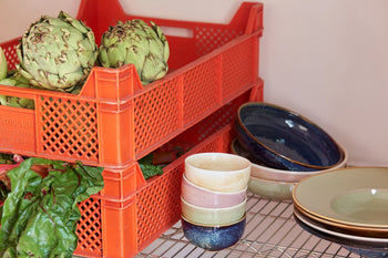 Two artichokes and leafy greens in an orange crate rest on a wire shelf beside stacked HKLIVING Chef Ceramics: Bowls and plates in assorted colors.