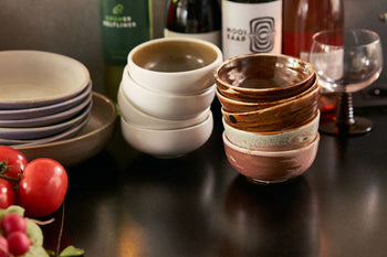 A stack of HKLIVING Chef Ceramics: Bowls by HKLIVING sits on a dark table with plates, fresh tomatoes, wine bottles, and an empty glass.