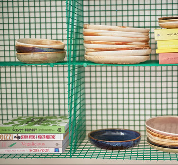 A green wire shelf displays HKLIVING Chef Ceramics: Deep Plate M and earthy-toned bowls, paired with colorful cookbooks. A matching green wire grid in the background creates a cohesive look.