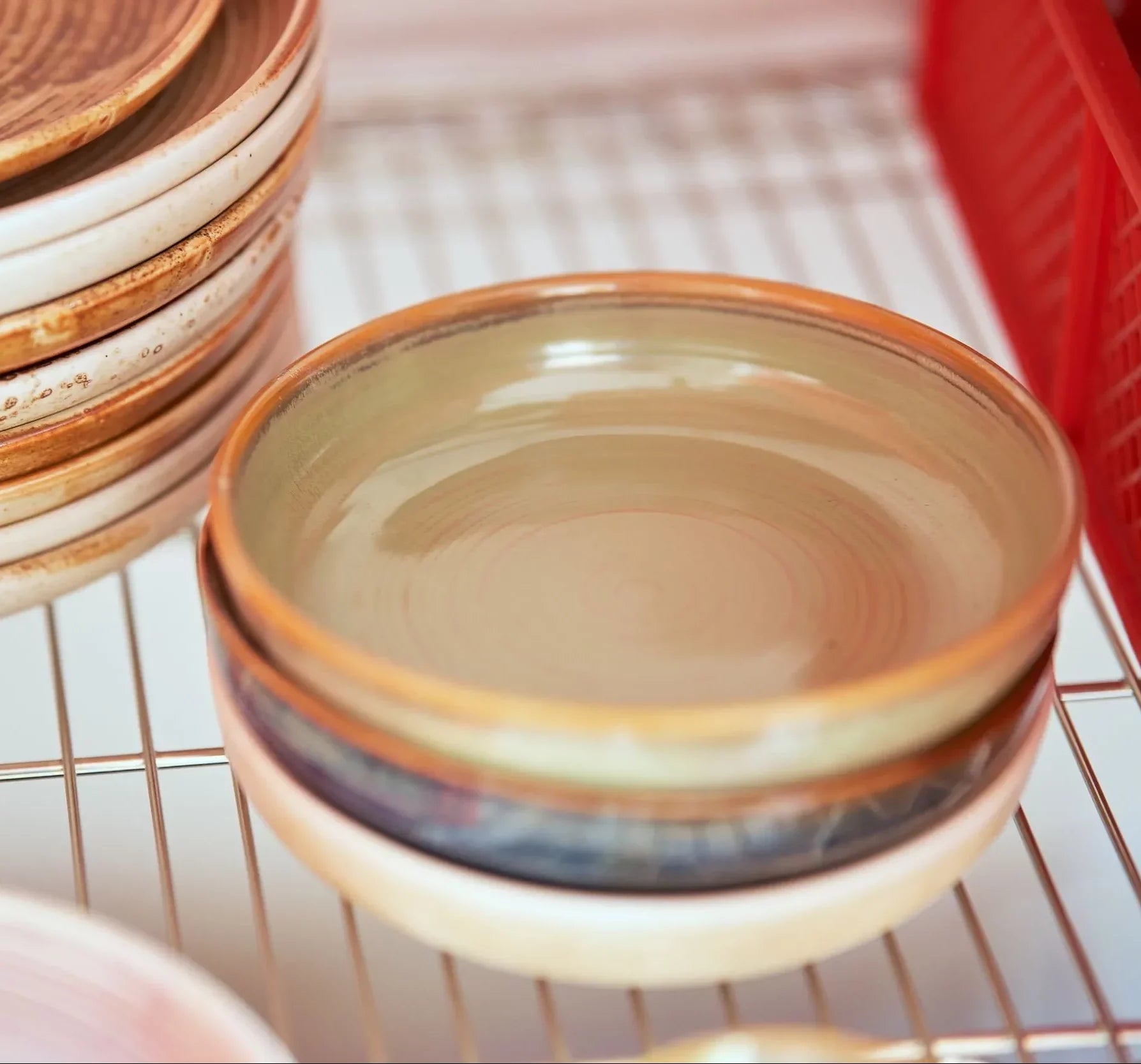 A stack of HKLIVING Chef Ceramics: Deep Plate M in earthy tones rests on a metal drying rack beside a red plastic container, with another taller stack of hand-glazed ceramic plates visible on the left.